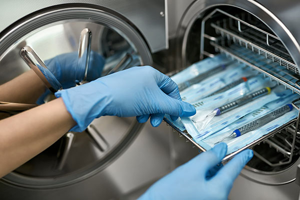 Dentist in blue latex gloves is taking a sealed dental probe from an autoclave sterilizer. Closeup horizontal photo.